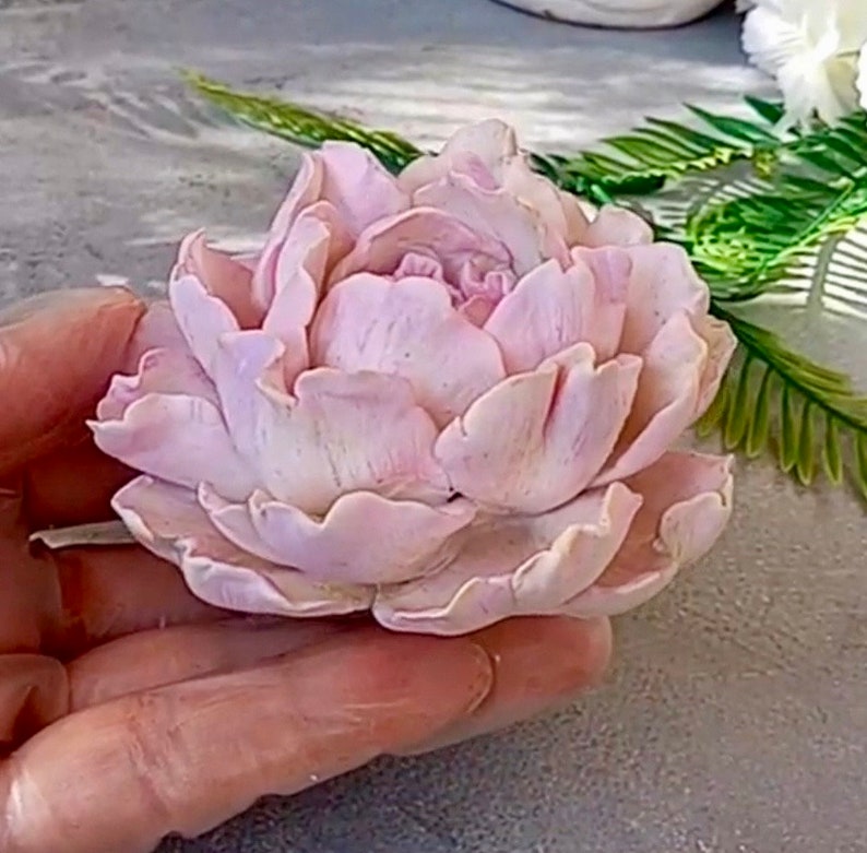 Pode incluir: Uma delicada flor de pe&ocirc;nia rosa feita &agrave; m&atilde;o com argila polim&eacute;rica. A flor tem uma apar&ecirc;ncia realista com detalhes intrincados e p&eacute;talas macias.