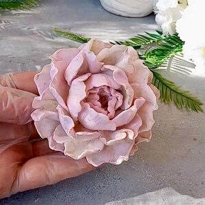 Pode incluir: Uma delicada flor de pe&ocirc;nia rosa feita &agrave; m&atilde;o com argila polim&eacute;rica. A flor tem uma apar&ecirc;ncia realista com detalhes intrincados e uma cor pastel suave.