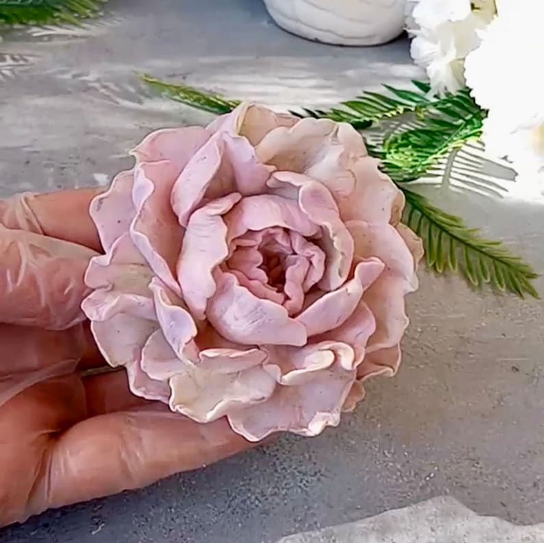 Pode incluir: Uma delicada flor de pe&ocirc;nia rosa feita &agrave; m&atilde;o com argila polim&eacute;rica. A flor tem uma apar&ecirc;ncia realista com detalhes intrincados e uma cor pastel suave.