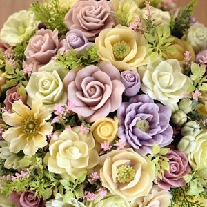 May include: A bouquet of artificial flowers in shades of pink, purple, and cream. The flowers are made to look like roses, carnations, and other types of blooms. The bouquet is arranged in a circular shape and is surrounded by green leaves.