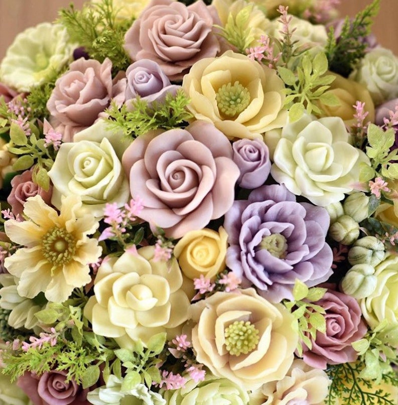 May include: A bouquet of artificial flowers in shades of pink, purple, and cream. The flowers are made to look like roses, carnations, and other types of blooms. The bouquet is arranged in a circular shape and is surrounded by green leaves.