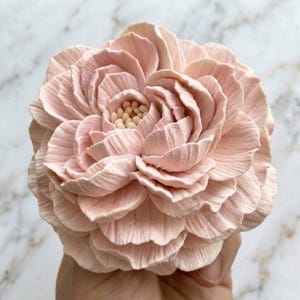 Op de afbeelding: Een lichtroze bloemensculptuur met talloze gelaagde, getextureerde bloemblaadjes in een cirkelvormig patroon. Het midden van de bloem bevat kleine, afgeronde, lichtgekleurde meeldraden. De bloemblaadjes hebben een matte afwerking en een realistische uitstraling.
