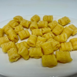 May include: A white plate filled with many golden-yellow, square cereal pieces. The cereal pieces have a slightly puffed appearance and a textured surface, suggesting a crispy texture. The lighting highlights the details of the food, which is a breakfast cereal.