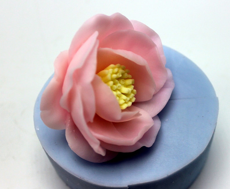 May include: A pink flower with a yellow center, sitting on a light blue surface. The flower is made of fondant or gum paste and is likely used for cake decorating.