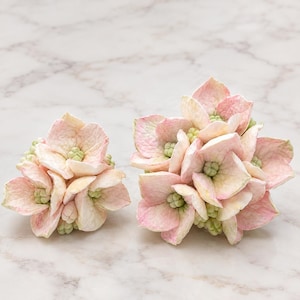 Könnte beinhalten: Zwei künstliche Hortensienblüten in zartem Rosa und Creme. Jeder Blütenkopf besteht aus mehreren geschichteten Blütenblättern mit hellgrünen Zentren, was ein realistisches botanisches Erscheinungsbild mit texturierten Oberflächen erzeugt.