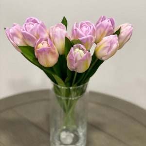 以下が含まれることがあります： 透明なガラスの花瓶に、薄い紫色のチューリップのブーケが活けられています。花瓶は灰色の木製のテーブルの上に置かれています。