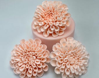 Può includere: Tre decorazioni a forma di fiore color pesca chiaro con petali arricciati, stratificati e complessi. Un fiore poggia su una base cilindrica liscia dello stesso colore, mentre due fiori identici sono posizionati davanti alla base.
