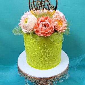 Pode incluir: Um bolo verde decorado com um desenho floral e um topo dourado de "Feliz Anivers&aacute;rio". O bolo est&aacute; em um suporte de bolo branco com uma base dourada.