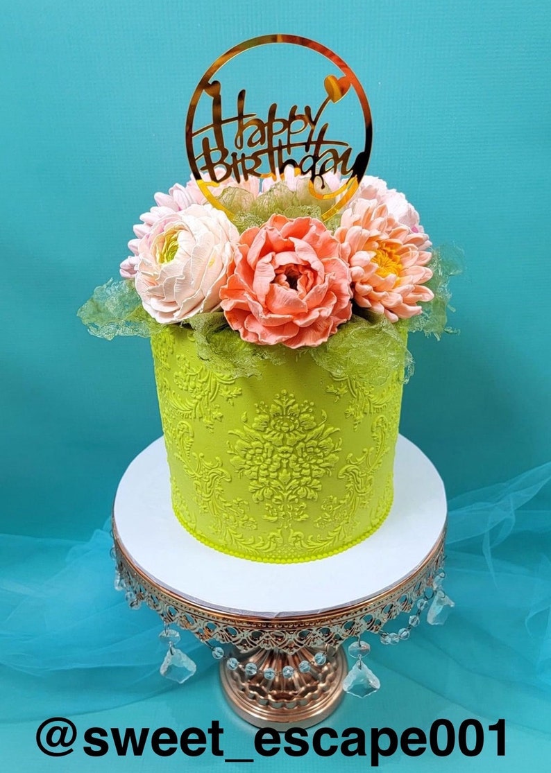 Pode incluir: Um bolo verde decorado com um desenho floral e um topo dourado de "Feliz Anivers&aacute;rio". O bolo est&aacute; em um suporte de bolo branco com uma base dourada.
