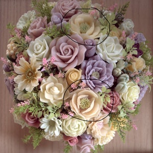 May include: A bouquet of artificial flowers made of soap. The bouquet is made up of various flowers in shades of pink, purple, white, and yellow. The flowers are arranged in a circular shape and are surrounded by green leaves.