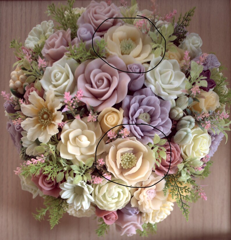 May include: A bouquet of artificial flowers made of soap. The bouquet is made up of various flowers in shades of pink, purple, white, and yellow. The flowers are arranged in a circular shape and are surrounded by green leaves.