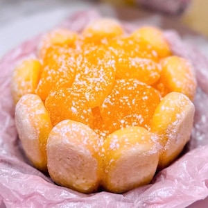 May include: A close-up of a dessert arrangement featuring bright orange, jelly-like candies dusted with powdered sugar. The candies are arranged in a circular shape, nestled in a pink paper container.