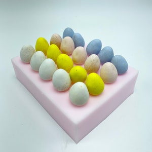 May include: A pink silicone mold with 18 small, colorful candy eggs in a grid pattern. The eggs are in shades of blue, yellow, and pink.