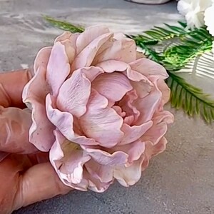 Pode incluir: Uma flor de pe&ocirc;nia delicada de cor rosa p&aacute;lido feita de argila polim&eacute;rica. A flor tem uma apar&ecirc;ncia realista com detalhes intrincados e p&eacute;talas macias e sobrepostas.