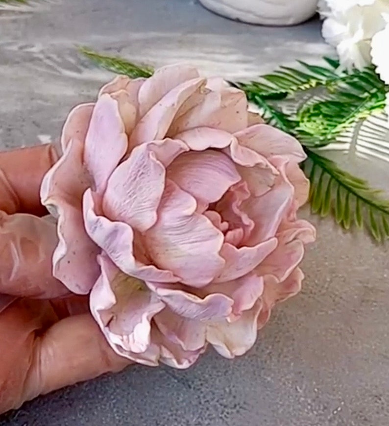 Pode incluir: Uma flor de pe&ocirc;nia delicada de cor rosa p&aacute;lido feita de argila polim&eacute;rica. A flor tem uma apar&ecirc;ncia realista com detalhes intrincados e p&eacute;talas macias e sobrepostas.