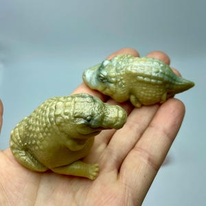 May include: Two small, detailed alligator figurines in shades of green and gold are held in a hand. The alligators have textured skin and are posed in a resting position. The background is a soft, neutral colour.