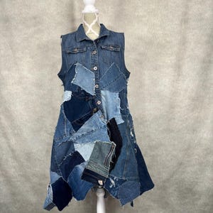 May include: A sleeveless denim dress with a patchwork design. The dress features a collared neckline, button-down front, and a mix of light and dark blue denim patches. The dress is displayed on a white mannequin.