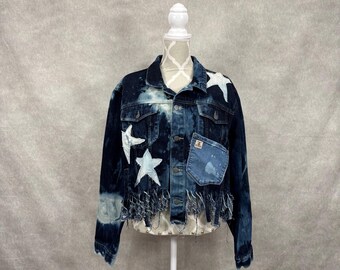 May include: A dark blue denim jacket featuring a bleached tie-dye effect, white star-shaped fabric appliqués, and a frayed fringe hemline. The jacket includes a chest pocket with a small rectangular label and a button-down front closure.