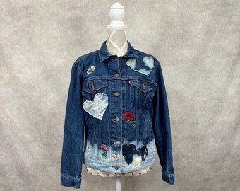 May include: A dark blue denim jacket featuring a light blue dip-dyed hem, metal button closures, two chest pockets, and various decorative patches including a white heart, a red flower, a pink flower, and a small daisy on the upper chest.