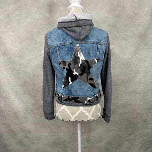 May include: A denim jacket with grey jersey sleeves and a matching hood. The back features a large camouflage star patch in shades of grey, black, and white, with a coordinating camouflage fabric band along the bottom hem of the denim panel.