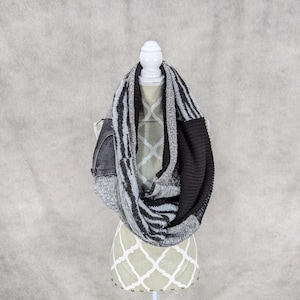 May include: A gray and black infinity scarf with a denim pocket detail. The scarf is made of a soft, knitted fabric and is perfect for adding a touch of warmth and style to any outfit.