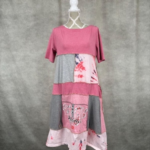 Op de afbeelding: Een roze en grijze patchworkjurk met een tie-dye patroon aan de onderkant. De jurk heeft korte mouwen en een ronde hals.