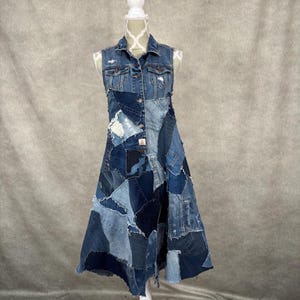 May include: A sleeveless denim dress displayed on a white mannequin. The dress is a patchwork of different shades of blue denim, featuring a button-down front and a flared skirt. The dress has a collar and is made from recycled jeans.