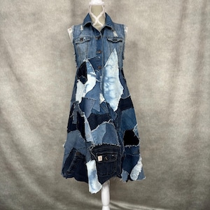 May include: A sleeveless denim dress on a white mannequin. The dress is a patchwork of various shades of blue denim, including light wash, dark wash, and bleached sections. It has a collared neckline and button-down front, with pockets visible on the skirt.
