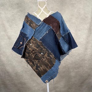 May include: A patchwork denim poncho with a V-neck design. The poncho features various shades of blue denim, brown camouflage fabric, and a brown embroidered patch. It has raw edges and pockets.