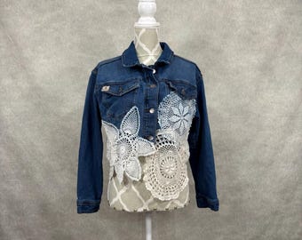 May include: A dark blue denim jacket featuring a button-up front, a collar, and long sleeves. The lower front section is embellished with several white and cream-coloured circular crochet doilies of varying sizes and intricate patterns.