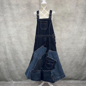 Può includere: Abito con bretelle in denim scuro con gonna patchwork. L'abito presenta spalline regolabili, una tasca a pettorina e un design unico della gonna realizzato con varie tonalità di denim blu. L'abito è esposto su un manichino.