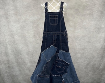 May include: Dark wash denim overall dress with a patchwork skirt. The dress features adjustable straps, a bib pocket, and a unique skirt design made from various shades of blue denim. The dress is displayed on a mannequin.