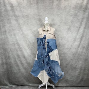 May include: A sleeveless beige button-down dress featuring an asymmetrical overlay of various blue denim fabric patches. The denim sections include visible pockets, seams, and frayed edges, layered over the solid beige fabric of the dress.