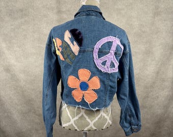 Peut inclure: Veste en jean bleue avec des écussons colorés, dont un symbole de paix, une fleur et des formes abstraites. La veste a un ourlet brut et court et est présentée sur un mannequin.