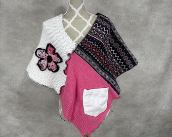 May include: A handmade poncho with a patchwork design. The poncho features a white knit section with a large flower applique, a pink section with a white pocket, and a patterned black and white section. The poncho is displayed on a mannequin.