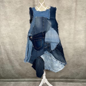 May include: A sleeveless denim dress made from patchwork denim fabric. The dress has a loose fit and a flowing silhouette. The dress is asymmetrical and has a raw hem.