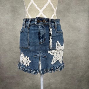 May include: A blue denim skirt with white crochet floral appliques and a frayed hem. The skirt has a button and zipper closure, front pockets, and a split in the front.