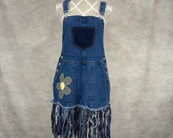 May include: A denim overall dress with a dark blue pocket and a flower appliqué. The dress has adjustable straps, button fastenings, and a fringed hem. The dress is displayed on a mannequin.