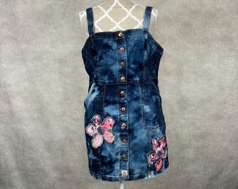 May include: A denim dress with a tie-dye pattern and pink floral embellishments. The dress has a button-up front and adjustable straps.