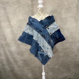 May include: A poncho constructed from repurposed blue denim fabric featuring various shades of indigo and light wash panels. The garment displays raw, frayed edges, visible metal buttons, and stitched seams arranged in an asymmetrical pattern.