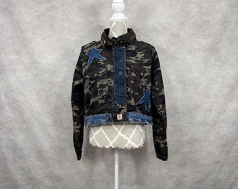 May include: A cropped jacket featuring a dark green, brown, and black camouflage pattern with two blue denim star patches on the chest. The jacket includes a dark blue denim button-up placket, a raw-edge denim hem, and two chest pockets with flaps.