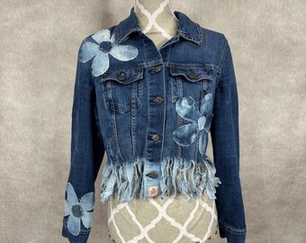 May include: A dark wash denim jacket with light blue floral appliques on the sleeves and chest. The jacket features a frayed hemline and button closures. The jacket is displayed on a mannequin.