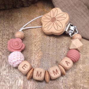 Könnte beinhalten: Ein Baby-Schnullerclip mit einem blumenförmigen Clip aus Holz. Der Clip hat eine Kette aus Holzperlen, eine rosafarbene Silikonperle in Rosenform, eine rosa Strickperle und Buchstabenperlen aus Holz mit der Aufschrift "EMMA".