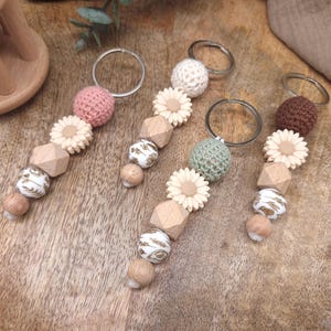 Peut inclure: Quatre porte-clés avec des perles décoratives. Chaque porte-clés comprend une boule crochetée, une fleur en bois, des perles géométriques en bois et des perles à motifs. Les porte-clés sont dans les tons de rose, blanc, vert et marron, avec des anneaux argentés.