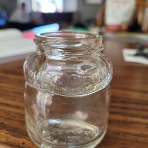 May include: A clear glass jar with a rounded body and a decorative band around the neck. The jar has a screw-top opening and is sitting on a wooden surface. The jar appears to be empty.