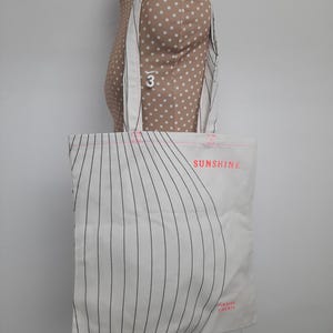 May include: White canvas tote bag with black stripes and the text "SUNSHINE" in pink. The bag has pink stitching and the text "GRAZIE CHERO" in pink at the bottom.