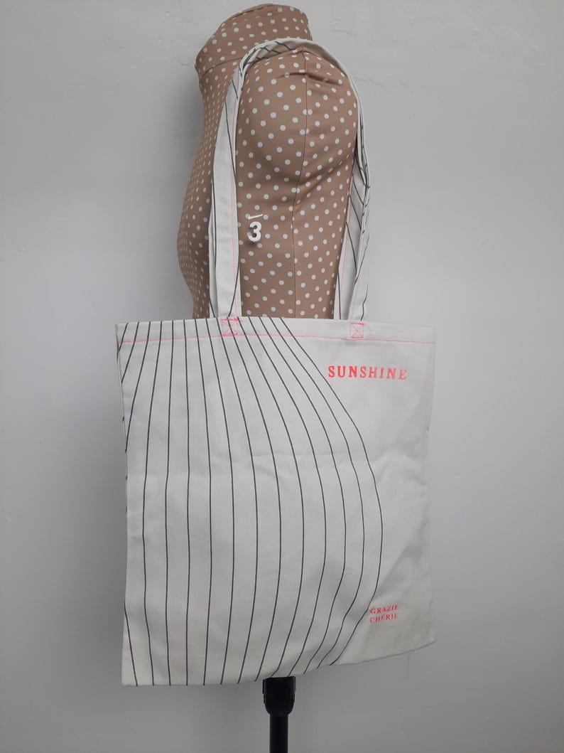 May include: White canvas tote bag with black stripes and the text "SUNSHINE" in pink. The bag has pink stitching and the text "GRAZIE CHERO" in pink at the bottom.