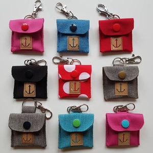 Könnte beinhalten: Neun kleine Stoffbeutel mit Ankermotiven und verschiedenen farbigen Knöpfen. Jeder Beutel hat einen silbernen Schlüsselring. Die Beutel sind in verschiedenen Farben, darunter rosa, rot, blau, grau und schwarz.