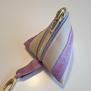 Könnte beinhalten: Beige dreieckige Tasche mit goldenem Reißverschluss und lila Band mit der Aufschrift "DO MORE OF WHAT MAKES YOU HAPPY". Die Tasche hat einen goldenen Verschluss und ein helllila Innenfutter.