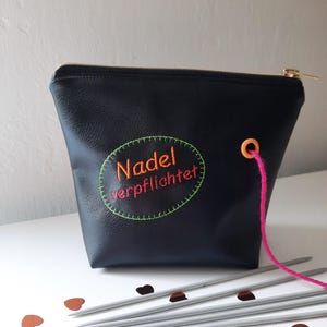 Könnte beinhalten: Schwarze Leder-Reißverschlusstasche mit einem grün und orange gestickten Kreis, auf dem "Nadel verpflichtet" steht. Die Tasche liegt auf einer weißen Oberfläche mit silbernen Stricknadeln und braunen herzförmigen Konfetti.
